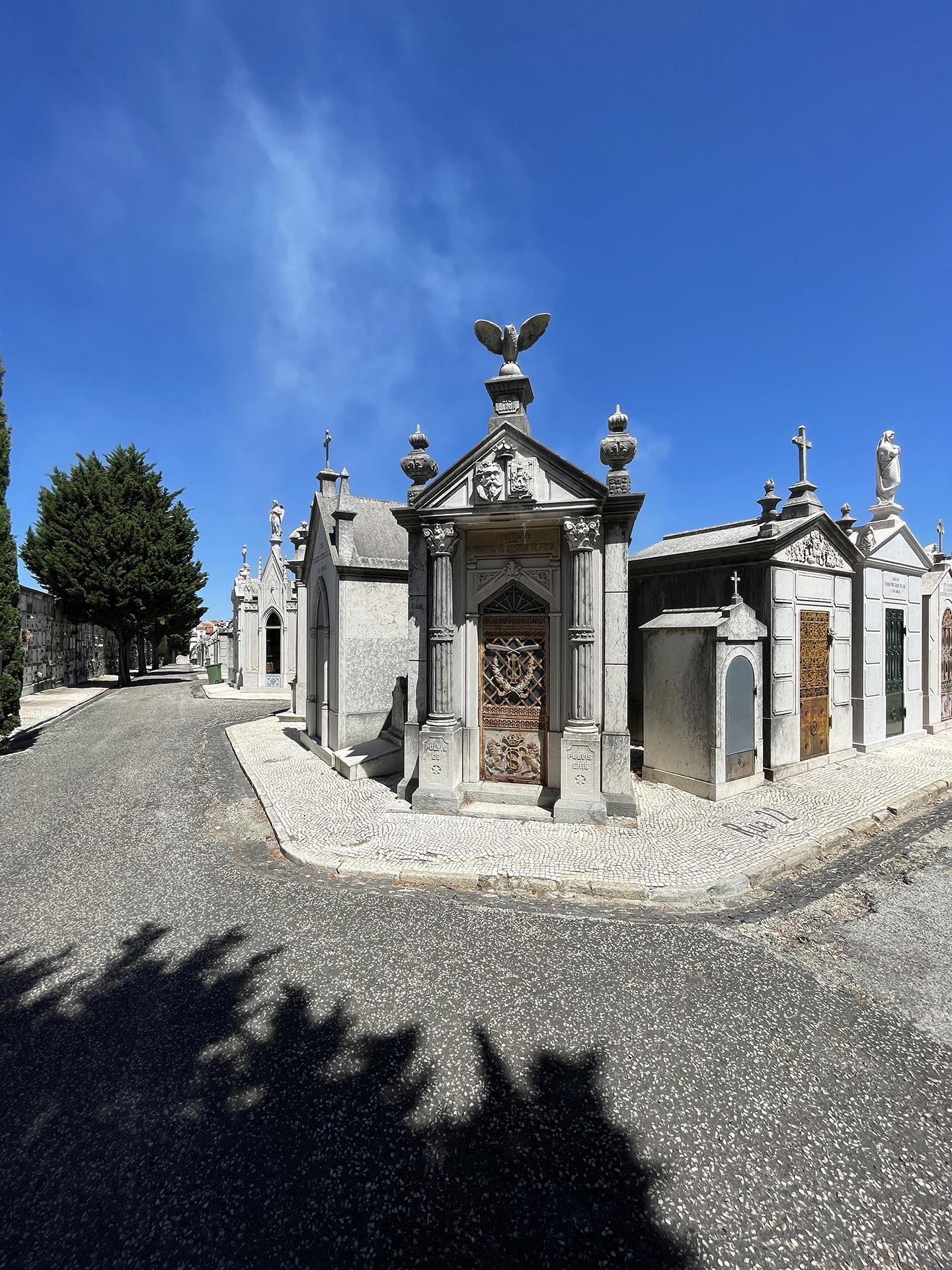 Photo: view of Alto de São João Cemetery. Mausoleum structures against a vivid blue sky, prominent stone carving of an owl on top of the corner structure.