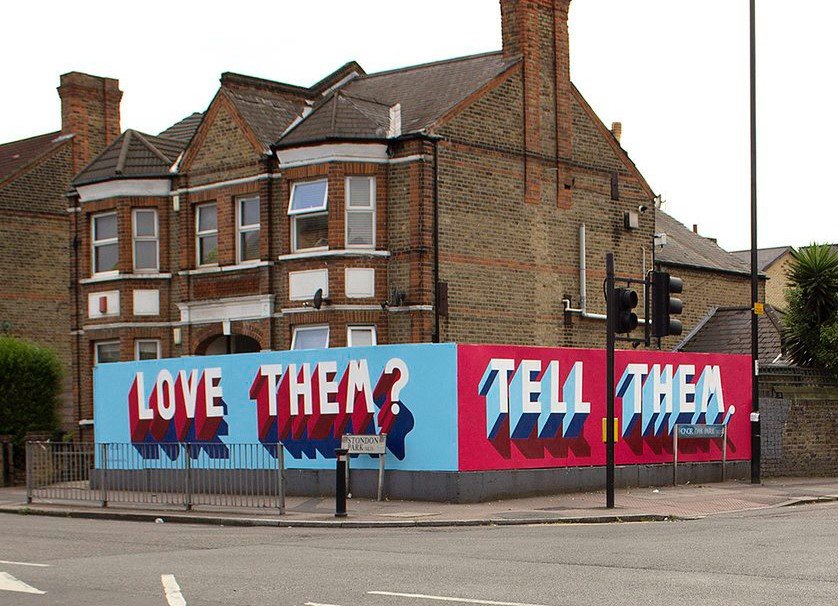 A mural wrapped arund a street corner with the words "Love Them?" written in white text with red shadow on a blue background on one side and "Tell Them." written in write text with a blue shadow ad red background on the other.