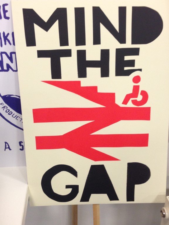 A white sign with black text "Mind the Gap" and a red symbol of the UK railway sign combined with a staircase. A hanidcaped icon at the bottom of the staircase shows that railways are not accessible