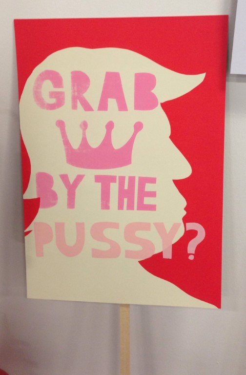 An anti-Trump protest poster depicting Trump's silhouette on a red background with the workds "Grad by the pussy?"